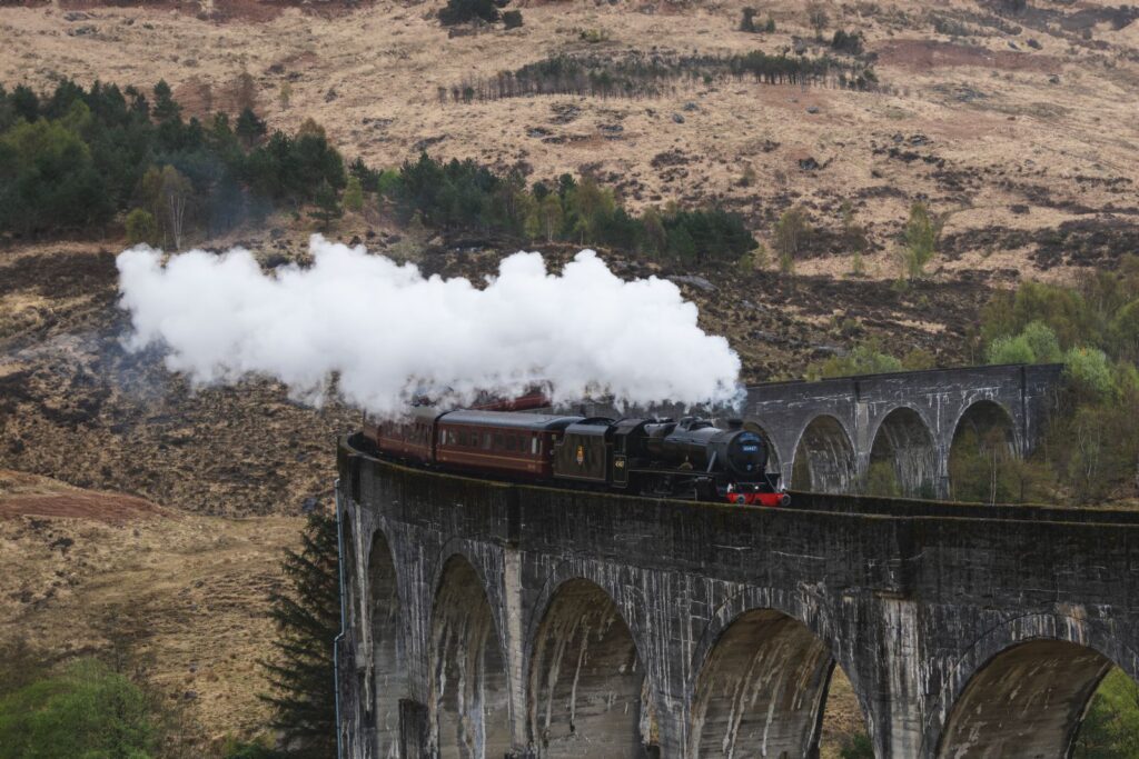 A steam locomotive and its carriages crossing the Glenfinnan Viaduct under a plume of white steam